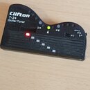 Černý plastový ladicí přístroj s nápisem Clifton T-24 Guitar Tuner. Na přední straně je displej, červená kontrolka a zelená LED dioda s označením b a…
