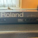 Detail zadní strany šedého elektronického hudebního zařízení s bílým nápisem Roland. Jsou zde vidět tři MIDI konektory označené jako THRU, OUT a IN.…
