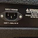 Detail zadní strany černého hudebního zesilovače s texturavaným povrchem. Je zde vidět napájecí konektor s nápisy INPUT POWER a 230V ~ 50Hz 95 W. Ved…