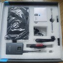 V bílém polystyrenovém packingu je umístěno několik předmětů. V dolní části je šedý přístroj s displejem a nápisem SENNHEISER XWIRELESS. Vedle něj je…