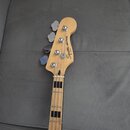 Fotografie zachycuje hlavu a část hmatníku basové kytary z přírodního světlého dřeva. Na hlavě je černý nápis „Squier JAZZ BASS“ a pod ním menší text…