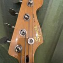 Fotografie zachycuje hlavu basové kytary z přírodního dřeva s lakovaným povrchem. Na hlavě jsou čtyři kovové ladičky a nápisy „Squier“ a „JAZZ BASS“,…