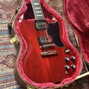 Červená elektrická kytara s tmavým hnedým hmatníkem a černým plechtrádem, umístěná v hnědém kufru s červenou výplní. Kytara má dva humbuckery, čtyři…