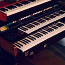 Fotografie zachycuje tři klavierní nástroje umístěné na stojanu nad sebou. Horní instrument má červené tělo, prostřední je černý s řadou barevných tl…