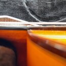 Detailní pohled na hranu smyčcového nástroje s lakovaným dřevěným povrchem v oranžovo-hnědém odstínu. Na nástroji je položen bílý smyček. Žádné značk…