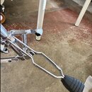 Detail chromovaného mechanismu bicího pedálu s černou gumovou podložkou pro nohu. Konstrukce je z kovových trubek a spojů, v horní části je vidět dal…