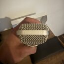 Fotografie zachicuje detail horní části mikrofonu s kovovou síťovkou stříbrné barvy. Kolem hlavice je umístěn hladký kovový prstenec v podobném odstí…