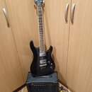 Černá elektrická kytara s dvojstupňovými humbuckery, opřená o černý kytarový kombo zesilovač. Na hlavě kytary je bílý nápis Cort. Zesilovač má na pře…
