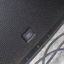 Detailní pohled na černý reproduktor s mřížkou z kovové síťky. Na mřížce je umístěn čtvercový černý štítek s bílým logem JBL. Vedle reproduktoru jsou…