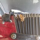 Detailní záběr na červený akordeon s černým méchem. Horní hrana méchu je výrazně poškozená a roztrhaná, s viditelným vnitřním materiálem. Povrch červ…