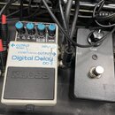 Dvě efektové pedály v černém kufru, propojené černými kabely. Levý pedál je bílý s modrým nápisem Digital Delay DD-2 a logem BOSS, má tři modré ovlád…