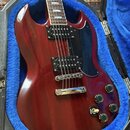 Kytara s červeným lakem a černým plechtrátem, umístěná v otevřeném kufru s modrou výplní. Nástroj má dva černé humbuckery, chromovaný most a čtyři če…