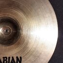 Detailní pohled na kovový činel zlatavé barvy s viditelným koncentrickým broušením povrchu. V dolní části je černé logo s nápisem ABIAN a grafickým s…