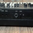 Zadní strana černého klavierního nástroje s řadou konektorů a ovládacích prvků. Jsou zde nápisy EXT INPUT, OUTPUT R, L/MONO, PHONES, DC IN, POWER, US…