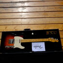 Elektrická kytara s tmelelovým povrchem sunburst a bílým pickguardem v černém tvrdém kufru. Na hlavě hmatníku je viditelné logo Fender. V kufru se na…