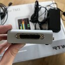Stříbrný kovový box s D-Sub konektorem na přední straně. Na krabici v pozadí je vidět nápis RTW a část textu for TM3. V pozadí jsou rovněž černý napá…