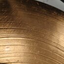 Detailní záběr na povrch mosazného činele s koncentrickými rýhami. Uprostřed je vyryt nápis PAISTE a níže menším písmem text MADE IN SWITZERLAND. Pov…