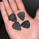 Čtyři černé kytarové trsátka v dlani. Každé má jiný povrch: jedno s otvory, dvě s jemným žebrováním a jedno s hvězdicovitým vzorem. Na trsátkách nejs…