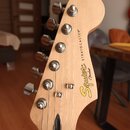 Fotografie zachycuje hlavu elektrokitary z přírodního světlého dřeva. Na povrchu je žluté logo Squier a černý nápis STRATOCASTER® s menším nápisem by…