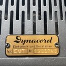 Detail zlatého kovového štítku připevněného na šedý perforovaný kovový povrch. Na štítku je nápis Dynacord Elektronik und Gerätebau, Typ: EMT a Nr. 1…