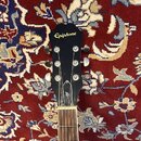 Fotografie zachycuje hlavu a část hmatníku elektrické kytary v černé barvě. Na hlavě je umístěno zlaté logo Epiphone a níže je vidět stylizované bílé…