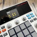 Bílý sampler s nápisem MPC SAMPLE. Zařízení má displej zobrazující vlnové průběhy, několik šedých otočných ovladačů označených K1, K2 a K3 a mřížku š…