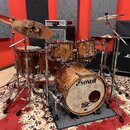 Bubnová sada s povrchem z hnědého dřeva a chromovaným kováním. Na basovém bubnu je bílý nápis SONOR a logo SQ2 DRUM SYSTEM. V pozadí jsou vidět Marsh…