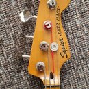 Fotografie zachycuje hlavu a horní část hmatníku basové kytary z přírodního dřeva. Na hlavě je černý nápis Squier JAZZ BASS a pod ním menší text BY F…