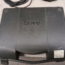 Černý plastový kufr s rukojetí. Na víku je vyembosované logo AKG a doprovodný symbol. V pravém dolním rohu je bílá nálepka s nápisy: Guitar Set, RF B…