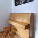 Světle hnědé pianino s lesklým povrchem a přívstupnou dřevěnou stoličkou. Nástroj je umístěn v rohu místnosti u bílé stěny, na které visí obraz s leo…