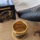 Zlatý kovový prsten (ligatura) s vyrytou značkou Jody Jazz. Uvnitř prstenu je černý nápis MT1. V pozadí je vidět černá krabička s nápisy SAVANNAH, NE…