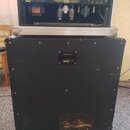 Fotografie zachycuje zadní stranu kytarového komba. Nahoře je zesilovač s nápisem MESA/BOOGIE a viditelnými elektronkami. Zesilovač stojí v hliníkové…
