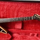 Elektrická kytara s přírodním dřevěným povrchem těla a černým hracím prknem. Na hlavě kytary je zlatý nápis Gibson a zlaté logo v podobu stylizedovan…