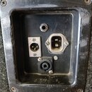 Fotografie detailu zadního panelu s konektory, osazeného do černého plastového rámu přišroubovaného k šedému texturovanému povrchu. Jsou zde vidět tř…