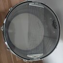 Fotografie znázorňuje snare drum s průhledným nylonovým popružem přes blanu. Na pravé straně blány je černé logo REMO a nápis STANDARD MADE IN USA. O…