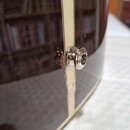 Detailní záběr na hranu tmavého, lesklého hudebního nástroje s bílým pruhem. Na hraně je umístěn kovový šroub s podložkou. V místě připojení šroubu k…