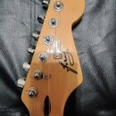 Fotografie zachycuje hlavu elektrické kytary z přírodního světlého dřeva s šesti chromovanými ladičkami. Na povrchu je umístěno černé logo "Fender".…