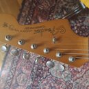 Fotografie zachycuje hlavu elektrické kytary z přírodního dřeva. Na povrchu je vidět logo Fender a nápis JAZZMASTER s doplňujícím textem WITH SYNCHRO…