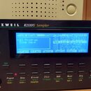 Černý sampler s nápisem KURZWEIL K2500RS Sampler. Přístroj má modrý LCD displej, na kterém je viditelný text ProgramMode, KB3 Program a Sine Wave. Po…