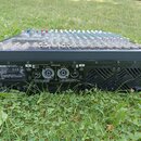 Černý mixážní pult s řadou posuvníků a knoflíků, umístěný na trávě. Na zadní straně je bílý nápis YAMAHA POWERED MIXER a další technické specifikace,…