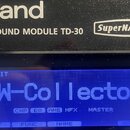 Detailní záběr na černý elektronický modul s bílými nápisy Roland, DRUM SOUND MODULE TD-30 a SuperNATURAL. Modul má modrý displej, na kterém je v hor…