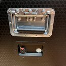 Detail kovové rukojeti s černým úchopem a připojovacího panelu na černém povrchu s hexagonálním vzorem. Na spodní hraně kovového rámu rukojete je mal…
