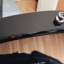 Detailní záběr na černý, pravděpodobně lakovaný obrubník hudebního nástroje. Na horní hraně je umístěn kovový šroub s maticí, který vykazuje známky o…
