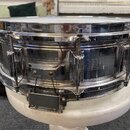 Chromovaná snare drum s kovovým korpusem. Na boku je vidět mechanismus napnutí pláště s dráty. Povrch vykazuje drobné škrábance a známky opotřebení.
