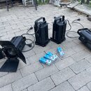 Čtyři černé reflektory s žebrovaným chladičem, z nichž dva stojí svisle a dva leží vodorovně. Jeden z ležících reflektorů má nasazené čtyři černé kla…