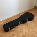 Černý tvrdý kufr na kytaru s kovovými zámky a rukojetí. Na povrchu je viditelné zlaté logo s nápisem "BANJO CASE". Kufr leží na dřevěné podlaze v ryb…