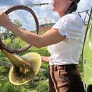 Žena v bílé blůzce a hnědých kalhotách hraje na mosazný nástroj s velkým zvonem. Tělo nástroje je obaleno tmavně hnědým materiálem s viditelným proší…