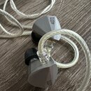 Šedé sluchátka typu in-ear s kovovým povrchem a černými silikonovými nástavky. Na obou sluchátkách je vyryt nápis KATØ MODERNO PRESENTS. Sluchátka js…