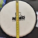 Kruhová bílá membrana bubnu s černým lemováním po obvodu. Uprostřed je černý nápis „NINO“ a pod ním text „PERCUSSION“. Přes nástroj je položen žlutýv…