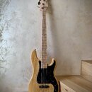 Elektrická basová kytara s přírodním dřevěným povrchem těla a hmatníku. Nástroj má černý pickguard s bílými tečkami, dva snímače a čtyři ladičky. Na…